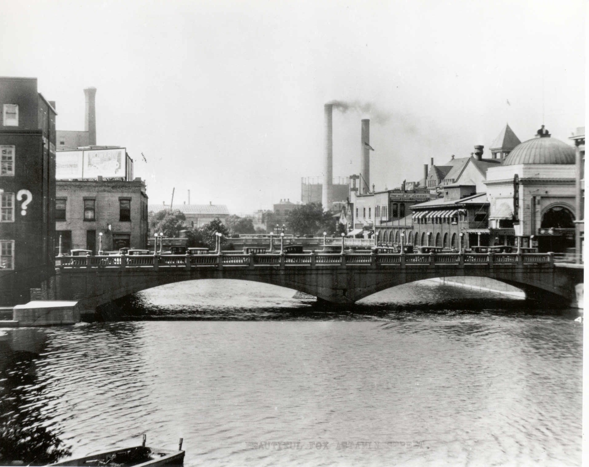A historic photo of a bridge in Aurora with a factory with two smoke stacks billowing smoke.